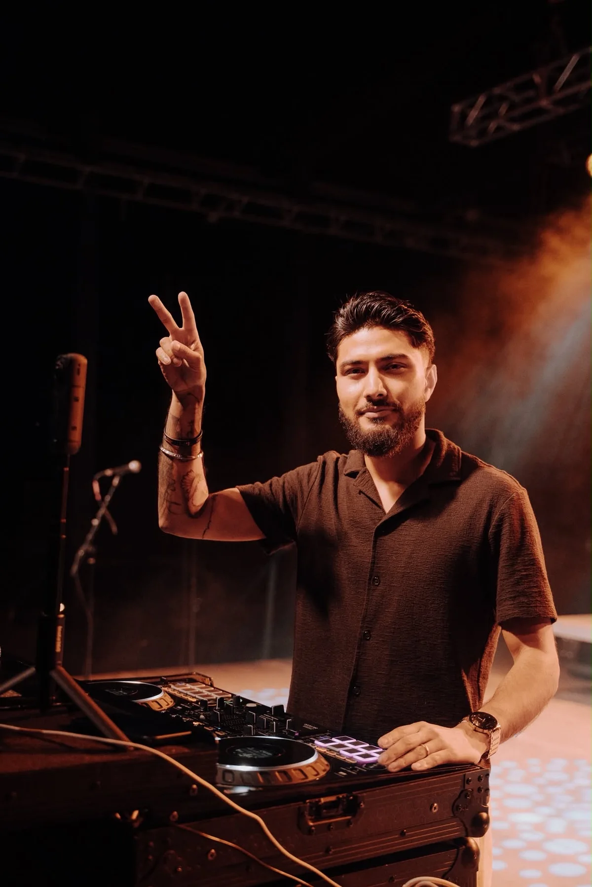DJ Sarif behind the decks, raising a peace sign under warm stage lighting